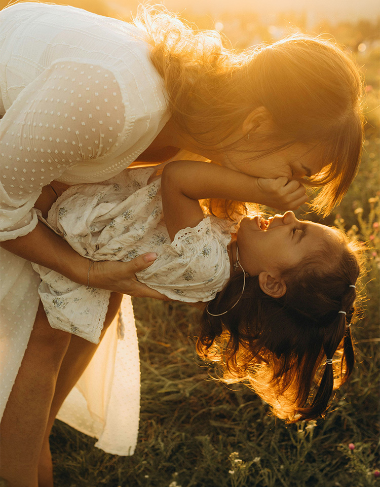 Mother embracing small daughter on a 90 degree angle in a grass field during sunset