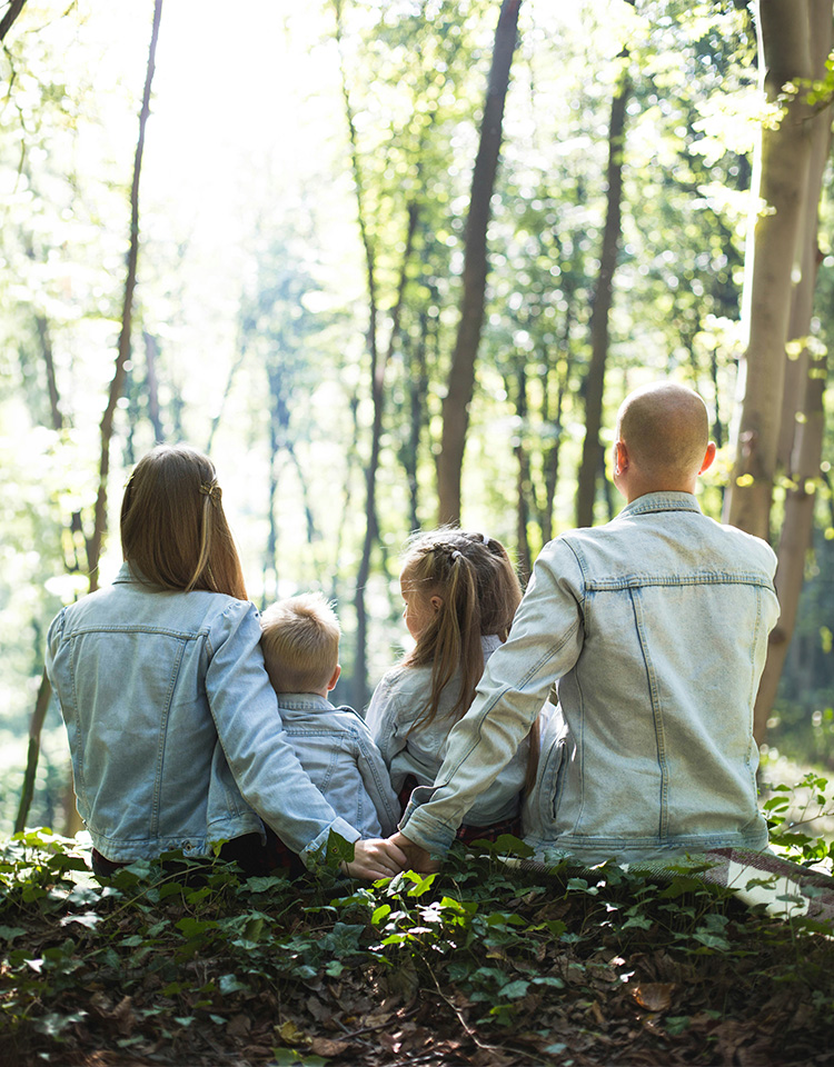 a family siting in a forested area with the husband and wife interlocking their fingers behind their two children back