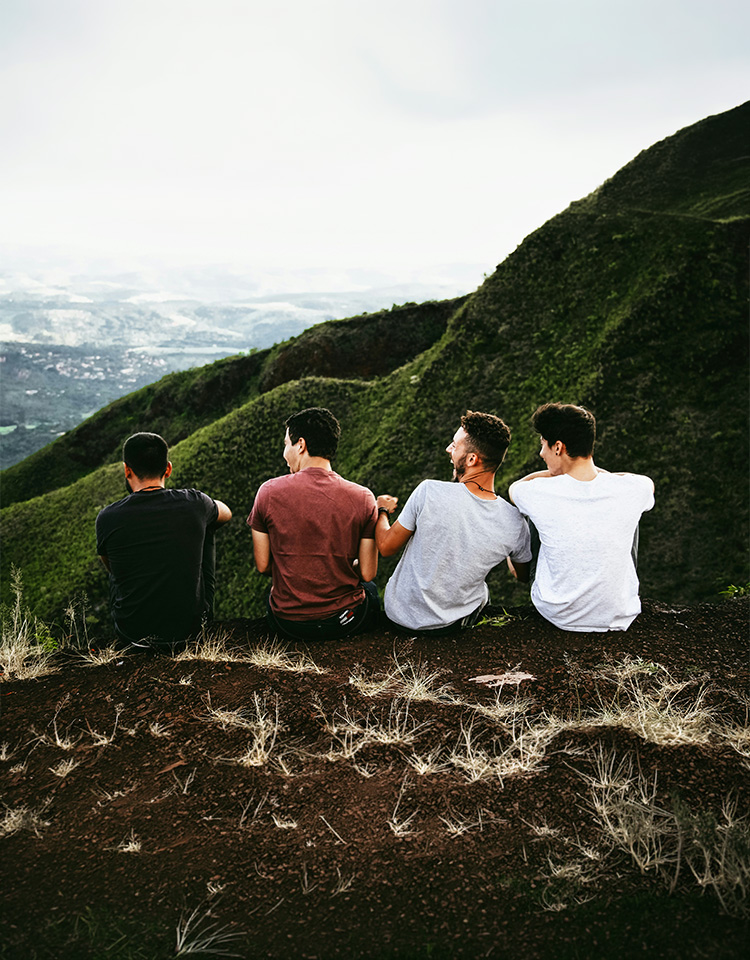 4 males sitting on the cliff of a mountain with their backs towards us overlooking the horizon