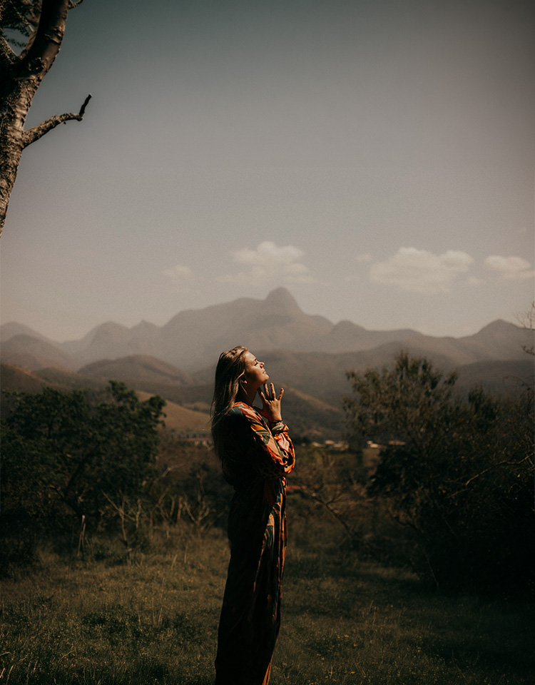 A woman who is standing in the mountainous area with her head upward, eyes closed and fingers interlocked to make a prayer hand.