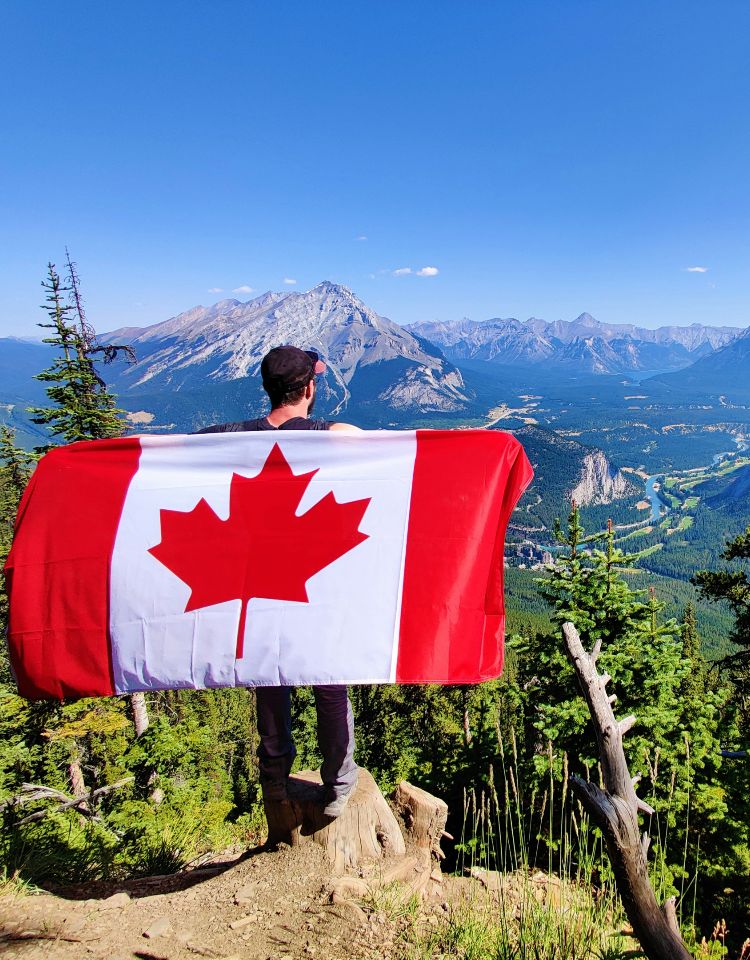 Man standing on mountain overlooking the northern scenery with a Canadian flag held out behind him like a cape.